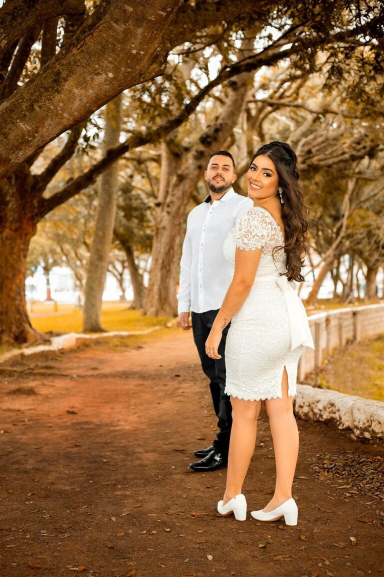 Casal sorridente em parque arborizado, mulher em vestido branco de renda, homem ao fundo em camisa azul clara, cenário romântico.